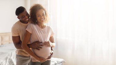 Pregnancy And Parenthood. Joyful Black Expectant Couple Embracing In Modern Bedroom Interior, Standing Together, Man Touching Wifes Growing Belly And Smiling. Panorama, Copy Space