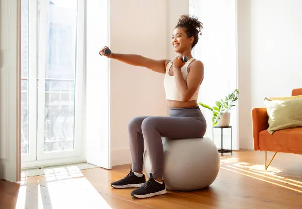 Sporty millennial lady with dumbbells perfecting her arm strength, exercising seated on stability ball, enjoys her morning workout routine in spacious living room interior. Empty space