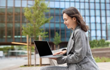 Young businesswoman browsing web on laptop sitting outdoors, representing IT and software career in urban street setting. Side view of lady websurfing on computer with empty screen mockup
