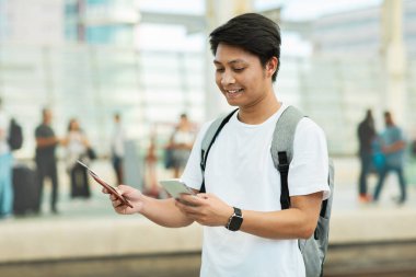 Portrait Of Young Asian Man Using Smartphone While Waiting At Airport, Happy Smiling Chinese Guy Checkin Online On Mobile Phone While Standing With Backpack At Crowded Terminal, Copy Space