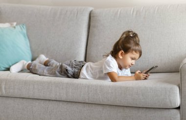 Cute toddler girl using smartphone while lying on comfortable sofa at home, little female kid playing games or watching cartoons, showcasing early engagement with technology in modern digital age