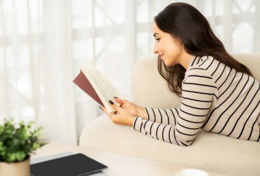 Smiling young caucasian lady read book, planning work, lie at sofa, enjoy free time, study, education in living room interior. Hobby and lifestyle cozy comfort at home