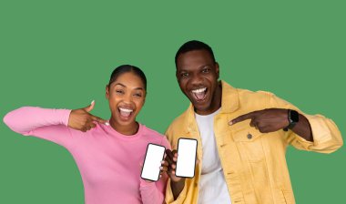 Cheerful Black Couple Pointing At Blank Smartphones And Showing Thumb Up, Happy African American Man And Woman Advertising New Mobile App Or Website, Posing On Green Background, Mockup