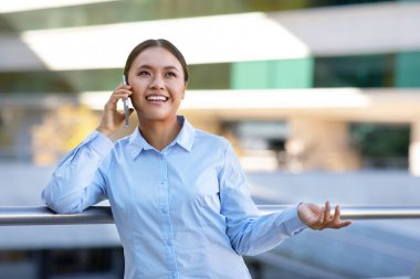 Happy Japanese entrepreneur woman chatting on smartphone, managing her business deals standing in urban area outside. Modern corporate and mobile communication concept