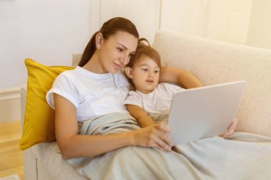 Mother and her little daughter with digital tablet relaxing under blanket on sofa, mom and preschool female child exploring online content together, watching cartoons or browsing internet