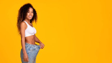 Perfect slim body. Smiling black woman wearing too large jeans proudly showing slimmed perfect body on yellow studio background. Healthy weight loss. Panorama, copy space