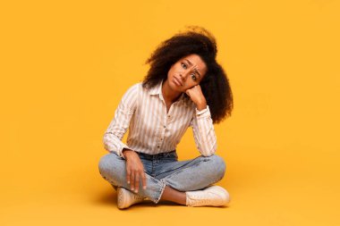 Contemplative young black woman with voluminous curly hair sits cross-legged on floor, hand resting on her cheek, deep in thought against vivid yellow background