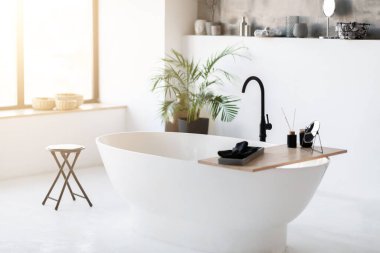 Spacious, modern bathroom interior featuring sleek freestanding bathtub in the middle, room filled with sunlight with minimalist luxury decor and potted plant in corner, copy space