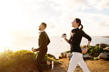 Energetic couple enjoying run along the coast, at sunny day, each carrying water bottles to stay hydrated, highlighting active lifestyle and fitness