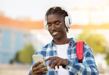 Cheerful young black man student using wireless headphones and smartphone outdoors, carrying his workbooks and backpack, enjoying favorite podcast on break at college, smiling at camera