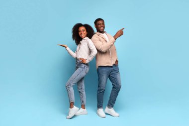 Cheerful young black man and woman standing back to back, smiling and pointing to their sides at free space against serene blue background