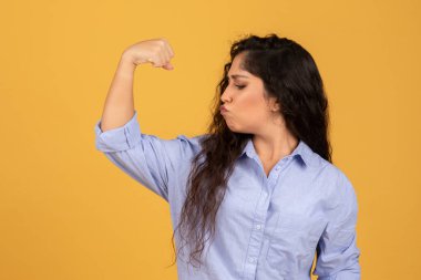Confident young woman with long dark hair flexing her bicep and puckering her lips, showcasing strength and empowerment on a solid yellow background, Female power, strong
