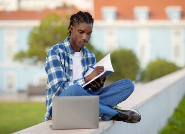 Focused Black young man student browsing on laptop and taking notes in workbook, while preparing for academic exam seated on campus park lawn outdoors, engaged in e-learning with computer