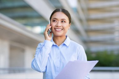 Young Chinese manager lady using her phone making business calls outdoors, standing with papers near corporate office center outside. Technology, communication and modern career