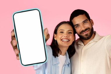 Beautiful cheerful cute multiracial young couple posing on pink background, happy millennial man and woman embracing and showing big phone with white blank screen, copy space, mockup. Offer for two