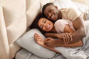 Loving black young man hugs his beautiful wife lying in bed in the morning. Happy couple embracing resting together in modern cozy bedroom interior. Copy space for text