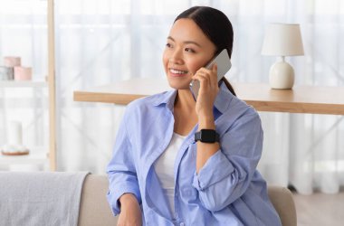 Portrait of cheerful young chinese lady have phone conversation at home, sit on couch in living room, talk with friend on smartphone and smiling, look at copy space. Cell operator, communication