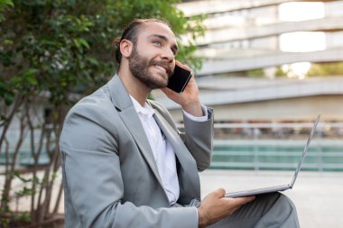 Happy young businessman talking on mobile phone and using laptop outdoors, millennial male entrepreneur embodying efficient communication and mobility, enjoying pleasant cellphone conversation