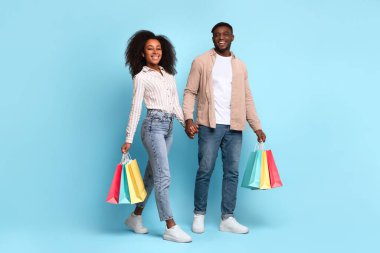 Happy young black couple holding hands and carrying colorful shopping bags, walking with relaxed, content smiles on vibrant blue background