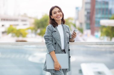 Young businesswoman smiling while texting on her phone, holding laptop computer standing outside in urban city area, browsing web and mobile applications on the way to work