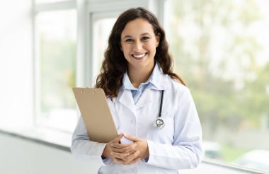 Cheerful millennial lady doctor in white coat, with stethoscope and clipboard standing next to window at clinic, hospital, copy space. Medical exam, health care consultation, prescription
