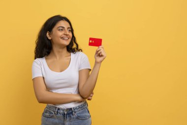 Positive smiling pretty young indian lady with red plastic credit card in her hand looking at copy space for advertisement, isolated on yellow studio background. Virtual banking, easy shopping