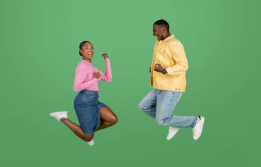 Emotional happy lucky young african american couple man and woman celebrating success together, jumping in the air, clenching fists and exclaiming, isolated on green studio background