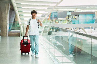Positive Asian Traveler Man Using Travel App On Smartphone, Walking By International Airport. Guy Passenger Booking Tickets In Mobile Application. Full Length, Copy Space