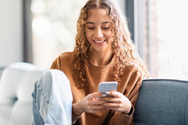 Teen girl websurfing on smartphone sitting on couch, spending time online and using mobile applications at home interior. Teenager lady immersed in digital world. Social media concept