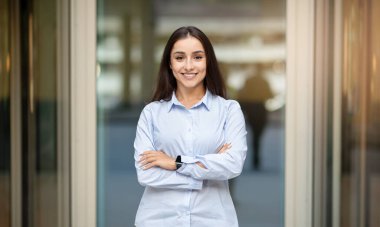 A confident happy pretty caucasian young woman manager with long hair, standing with crossed arms, wearing a light blue shirt and a smartwatch, smiling in an office corridor