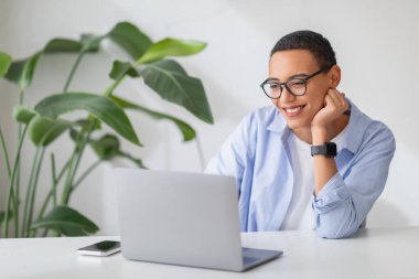 Cheerful latin young woman student with glasses looking at her laptop screen, leaning on her hand, wearing a casual blue shirt and a smartwatch in a fresh, plant-filled room