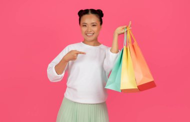 Shopping Sales. Smiling Asian Woman Pointing At Paper Shopper Bags In Her Hand, Happy Korean Female Demonstrating Purchases, Standing Isolated On Pink Studio Background, Copy Space