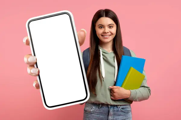 Online Education, Mobile App. Pretty Teen Girl Posing With Workbooks Over Pink Studio Background, Happy Student Wearing Backpack Showing Phone With White Blank Screen, Copy Space, Mockup