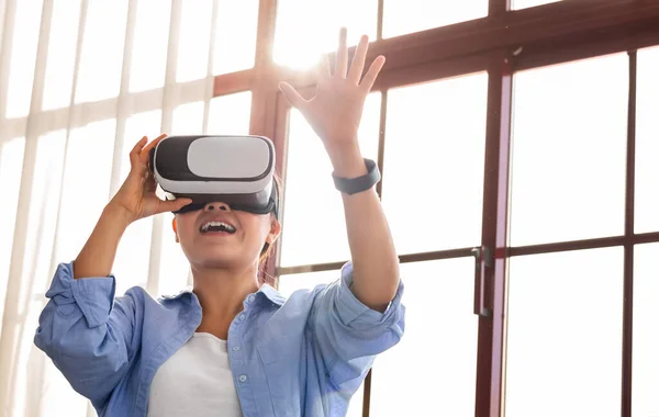 Virtual entertainments concept. Portrait of happy young asian woman in VR headset standing by window at home, playing interactive video games, exploring augmented reality, sun flare
