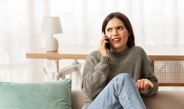 Communication problems. Young irritated hispanic woman arguing with interlocutor via cellphone, having quarrel with client or boyfriend, sitting at home, looking at copy space, gesturing