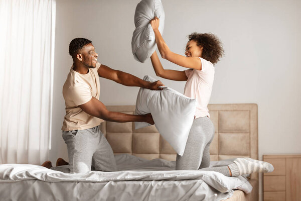 Young black couple playfully engages in a pillow fight, laughing with joy while having fun together on carefree morning, in modern home bedroom, fighting holding pillows