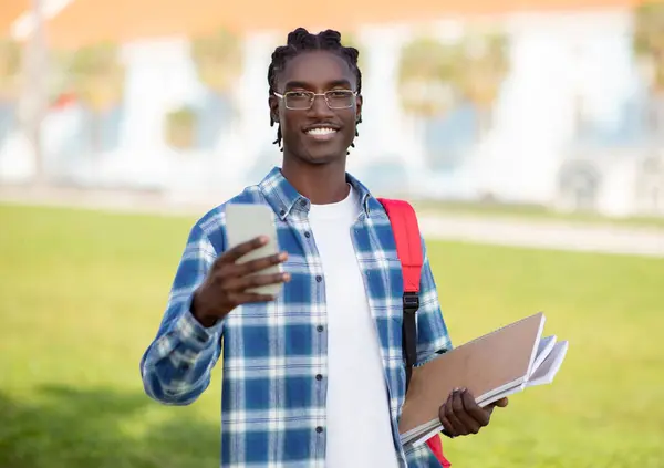 Happy Black Student Guy Holds Smartphone And Workbooks Standing Outside, Smiling To Camera While Texting And Scrolling Social Media Applications, Enjoying Study Break Outdoors College Campus