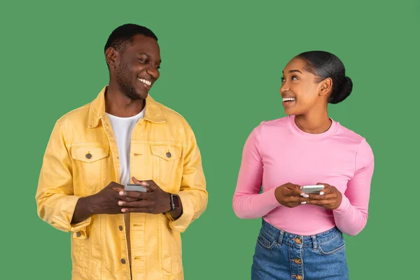 Cheerful millennial african american lady and guy typing on phone, chatting at gadget, isolated on green studio background, looking at each other and smiling. Shopping, communication online app