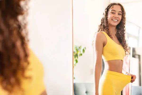 Happy fit teen girl looking at reflection in mirror and pulling fitness pants waist showing result of dieting and slimming, motivated after workout training at home. Sport and weight loss