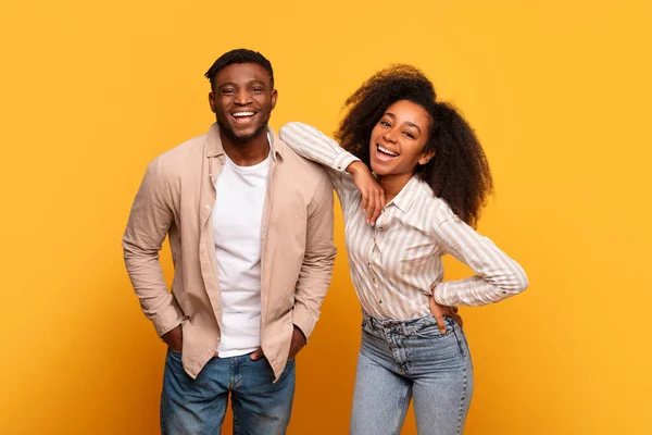 Joyful young black man and woman share laugh, leaning on each other with bright smiles, dressed casually in shirts and jeans against sunny yellow backdrop