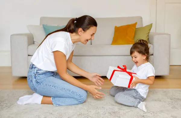 Excited little daughter receiving gift from mom at home, happy cute preschool female child getting birthday present box with red ribbon, sitting with mommy on floor in living room interior
