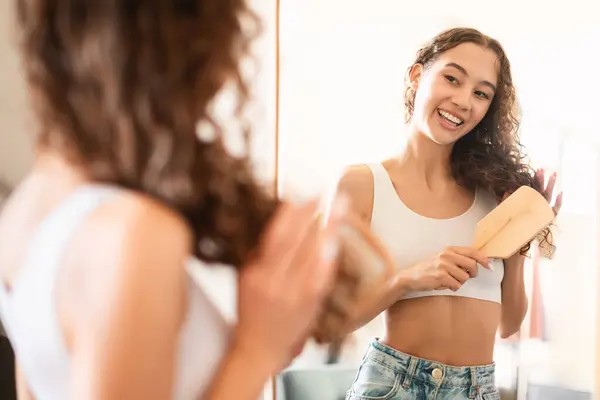 Happy Teen Girl Brushing Hair With Wooden Hairbrush, Smiling To Her Reflection In Mirror At Home, Selective Focus. Young Lady Enjoying Her Daily Haircare Routine In The Morning