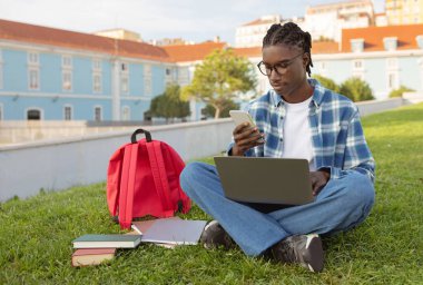 E-learning application. African American university student guy with laptop scrolling through phone sitting outside, browsing websites for educational information, studying online