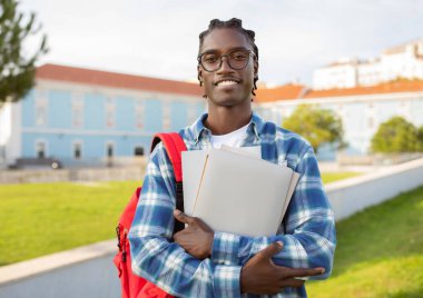 Cheerful black college student guy stands with his laptop, backpack and books smiling to camera at university park outside, wearing glasses. Education and academic success concept