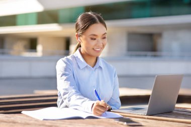 Korean business lady near laptop browsing and writing, sitting outdoors urban office center, juggling e-learning and professional tasks with modern online technology