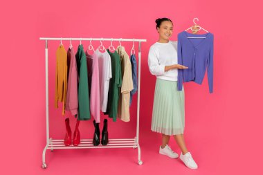 Happy asian female showing new blouse while standing near clothing rack, smiling korean shopper lady exploring different styles, posing isolated on pink studio background, full length shot