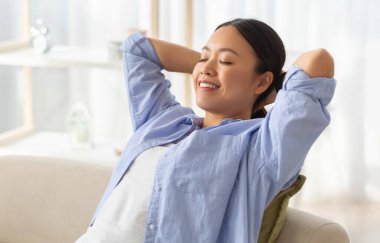 Overjoyed happy young asian woman wearing comfy clothing relaxing on couch at home, smiling with closed eyes, holding arms behind head, enjoying weekend, dreaming, copy space