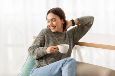 Overjoyed attractive brunette young woman wearing casual clothing relaxing on couch at home, holding white mug, smiling and stretching her body, drinking coffee at weekend, copy space