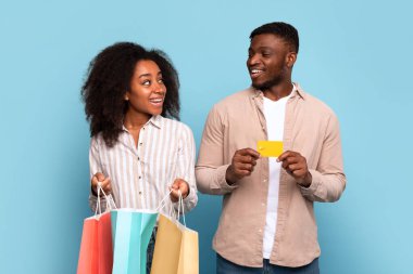 Excited young black couple holding colorful shopping bags and credit card, looking at each other with joy against blue background