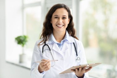 Cheerful attractive brunette young european woman doctor wearing medical uniform white coat and stethoscope posing at clinic or hospital next to window, holding clipboard, smiling at camera